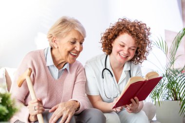 Bakıcı kadın huzurevinde mutlu bir yaşlı kadınla otururken kitap okuyor. Mutlu yaşlı kadın beyaz koltukta oturuyor ve hemşireyi yüksek sesle kitap okurken dinliyor.