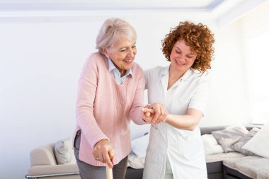 Young carer supporting senior disabled woman with walking stick. Portrait of happy female caregiver and senior woman walking together at home. Professional caregiver taking care of elderly woman.