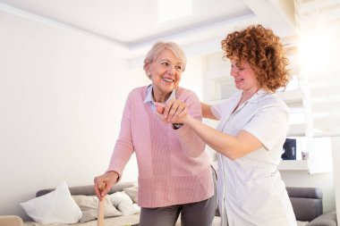 Smiling nurse helping senior lady to walk around the nursing home. Portrait of happy female caregiver and senior woman walking together at home. Professional caregiver taking care of elderly woman.