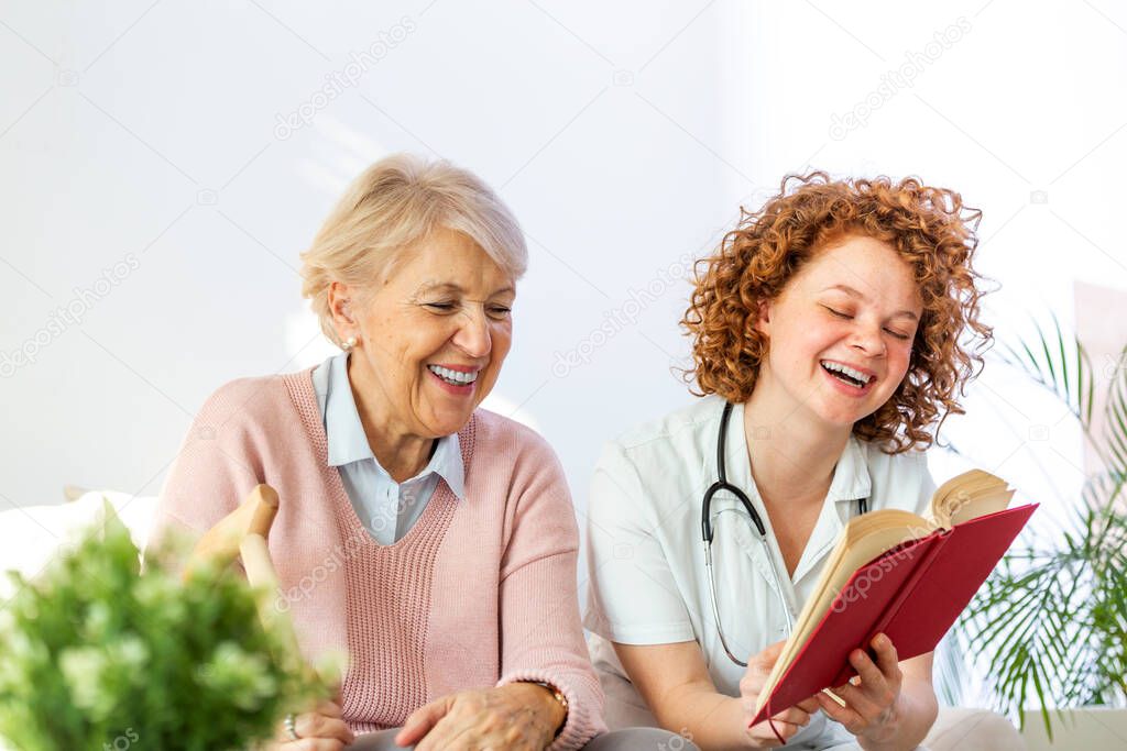 Woman caregiver reading a book while sitting with happy senior woman at nursing home. Happy elder woman sitting on white sofa and listening to nurse reading a book out loud
