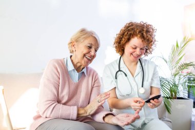 Happy senior woman having her blood sugar measured in a nursing home by her caregiver. Happy nurse measuring blood sugar of a senior woman in living room - diabetes and glicemia concept