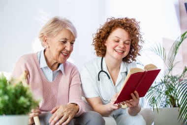 Woman caregiver reading a book while sitting with happy senior woman at nursing home. Happy elder woman sitting on white sofa and listening to nurse reading a book out loud