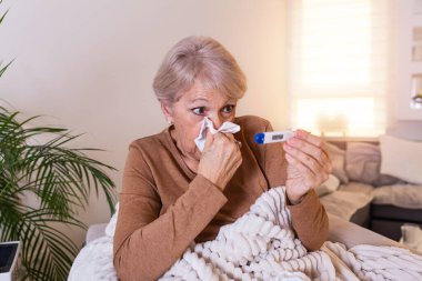 Hastalık, mevsimsel virüs problemi konsepti. Yaşlı kadın kanepede yatıp termometredeki sıcaklığa bakarak hasta oluyor. Hasta yaşlı kadın yüksek ateşle yatakta yatıyor. Soğuk algınlığı ve migren.