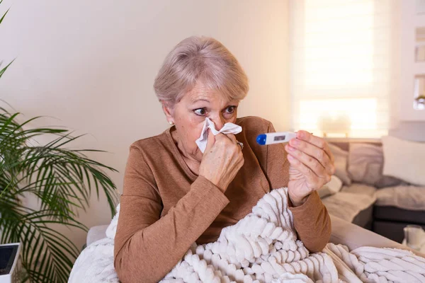 Hastalık, mevsimsel virüs problemi konsepti. Yaşlı kadın kanepede yatıp termometredeki sıcaklığa bakarak hasta oluyor. Hasta yaşlı kadın yüksek ateşle yatakta yatıyor. Soğuk algınlığı ve migren.