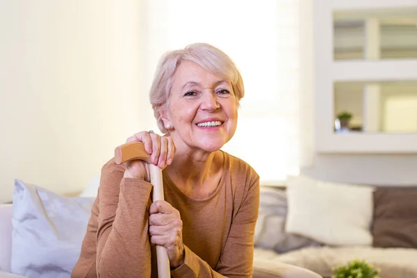 Smiling grandmother sitting on couch. Portrait of a beautiful smiling senior woman with walking cane on light background at home. Old woman sitting with her hands on a cane