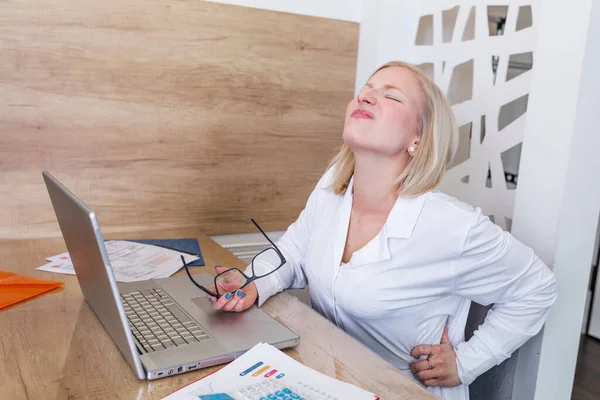 Portrait of young stressed woman sitting at home office desk in front of laptop, touching aching neck with pained expression, suffering from neck pain after working on pc