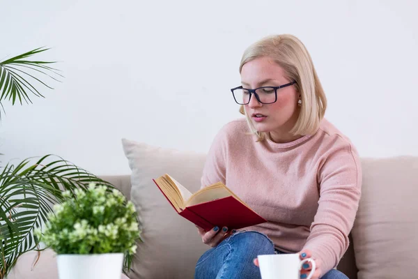 Young woman at home sitting on modern sofa relaxing in her living room reading book and drinking coffee or tea. White cozy bed and a beautiful girl, reading a book, concepts of home and comfort