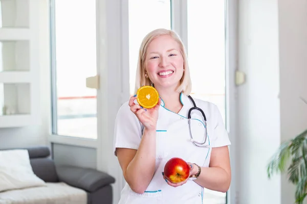 Smiling nutritionist in her office, she is holding a fruit and showing healthy vegetables and fruits, healthcare and diet concept. Female nutritionist with fruits