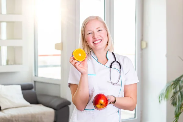 Smiling nutritionist in her office, she is holding a fruit and showing healthy vegetables and fruits, healthcare and diet concept. Female nutritionist with fruits