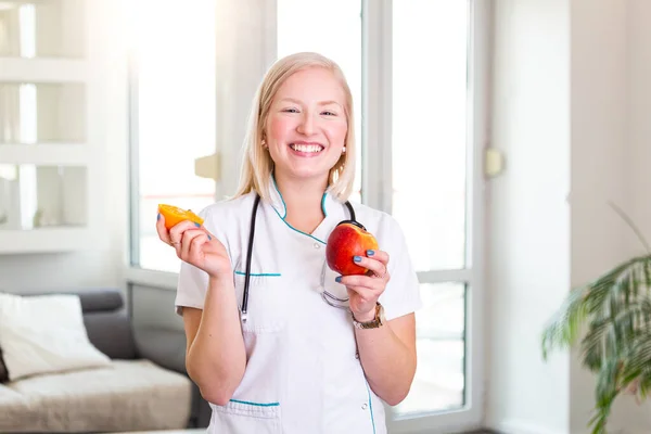 Smiling nutritionist in her office, she is holding a fruit and showing healthy vegetables and fruits, healthcare and diet concept. Female nutritionist with fruits