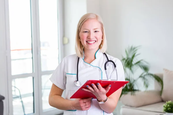 Female doctor holding clip board and smiling. Woman doctor standing straight in home care hospital. Close-up of a female doctor using clip board with stethoscope around her neck