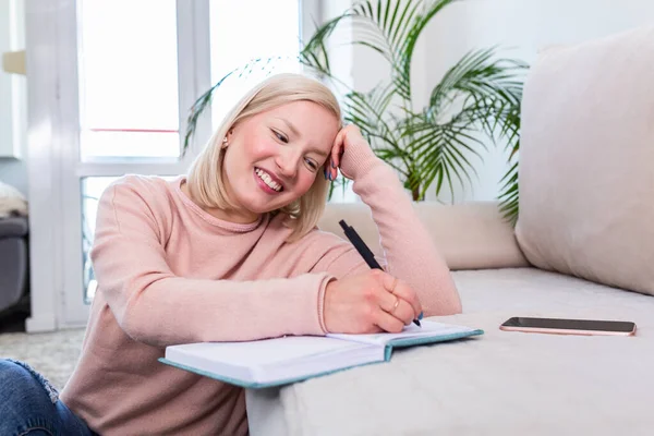 Girl studying the day before the exam, writing notes with a pencil. Pretty young girl doing her studies at home sitting on the floor in the living room with a book reading a binder of class notes