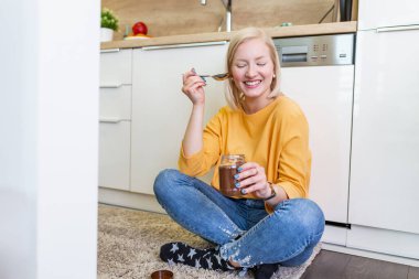 Güzel genç albino kız gündelik giysiler içinde ev mutfağında ahşap zeminde otururken lezzetli çikolata tadı alıyor..