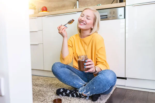 Young woman eating chocolate from a jar while sitting on the wooden kitchen floor. Cute albino girl indulging cheeky face eating chocolate spread from jar using spoon savoring every mouthful