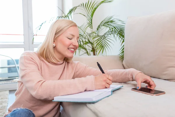 Girl studying the day before the exam, writing notes with a pencil. Pretty young girl doing her studies at home sitting on the floor in the living room with a book reading a binder of class notes