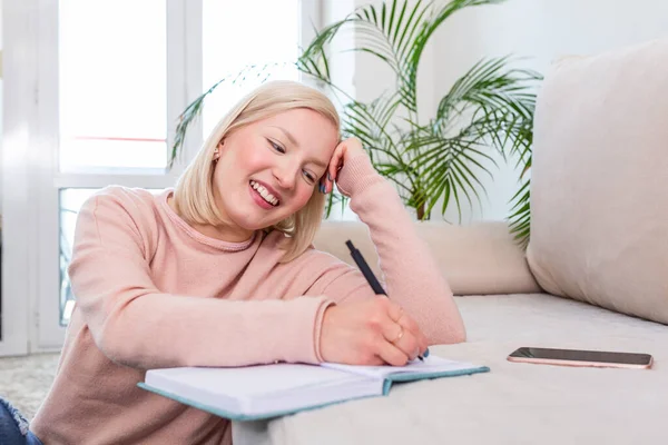 Girl studying the day before the exam, writing notes with a pencil. Pretty young girl doing her studies at home sitting on the floor in the living room with a book reading a binder of class notes