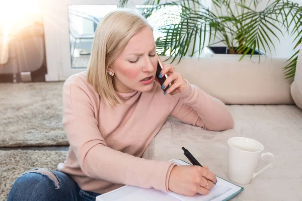 Girl working from home, talking on the phone and writing notes in her note book. Entrepreneur or student working or studying at home and writing notes, working from home