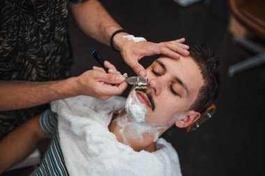 Man mith mustaches having a shave. Razor in hands of specialist barber. Young man getting an old-fashioned shave at the barber shop. Closeup, retro styled imagery