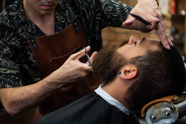 Bearded male sitting in an armchair in a barber shop while hairdresser trip his beard with