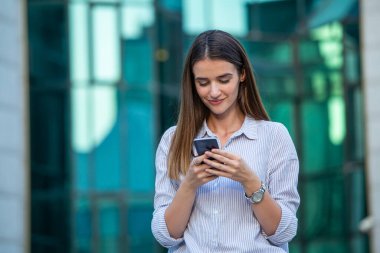 Young student is browsing on her pda while having a spring walk in the town outside, in a formal outfit, smiling, going to meet friend