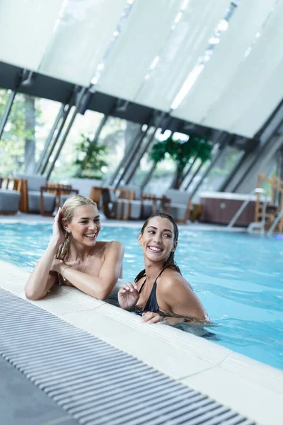 Shot of two women cheering with drinks near swim pool. Red, green and yellow different cold cocktails with ice cubes, straws, transparent clean blue aqua in pool