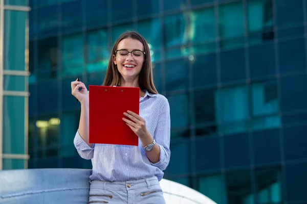 Happy beautiful business woman with clipboard. Portrait Of Successful Business Woman Holding clipboard. Photo business woman wearing suit and holding documents in hands. Panoramic windows background.