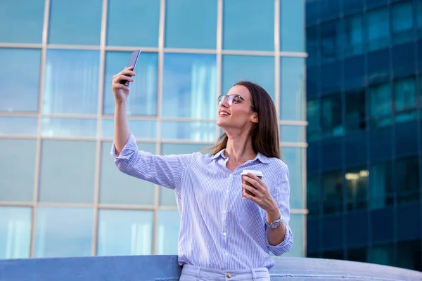 Executive business woman looking at mobile smartphone and drinking coffee from disposable paper cup in the street with office buildings in the background.