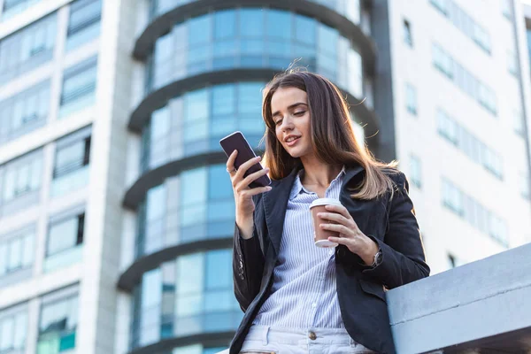 confident businesswoman texting on her mobile phone while drinking coffe in front of glass window of skyscraper building dreaming of success thinking of future business vision and new goals