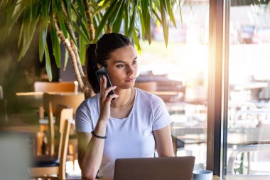 Kahve fincanı ve dizüstü bilgisayarlı genç bir iş kadını akıllı cep telefonuyla konuşuyor. Bir kadın mola veriyor ya da serbest çalışıyor. Kahve dükkanından iş yapmak.