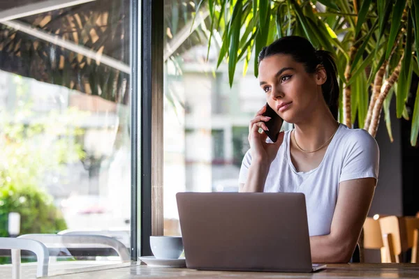 Elinde kahve fincanı ve laptopuyla akıllı telefonuyla konuşan genç bir kadın. Bir kadın mola veriyor. Kahve dükkanından iş yapmak.
