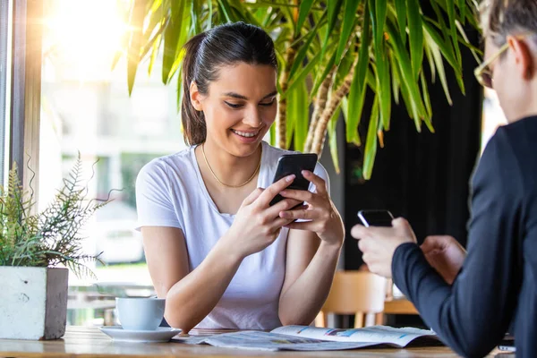 One-on-one meeting.Two young business women sitting at table in cafe. Girl shows colleague information on laptop screen. Girl using smartphone, blogging. Teamwork, business meeting. Freelancers working.