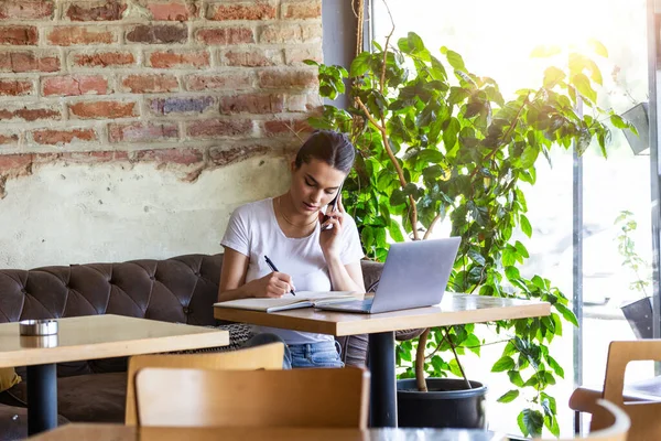 Young business woman with coffee cup and laptop talking on her smart mobile phone. Woman taking a break or freelancing. Doing Business From the coffee shop
