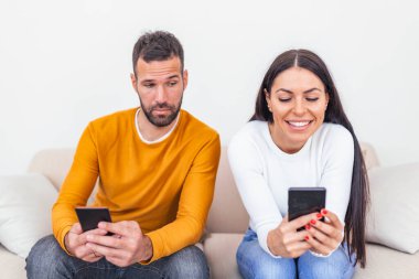 Kıskanç erkek arkadaş, kız arkadaşı üzgün üzgün bakarken onun telefonunu izliyor. Evde akıllı telefon kullanan genç bir çift. Kıskanç erkek arkadaş kız arkadaşının telefonunu gözetliyor..
