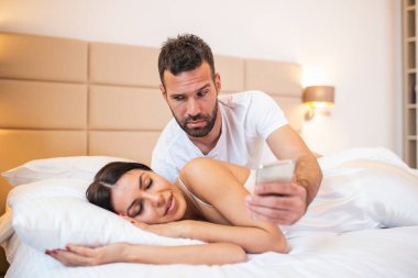 Jealous husband spying the phone of his partner while she is sleeping in a bed at home. Shocked jealous husband spying the phone of his wife while she sleeping in bed at home