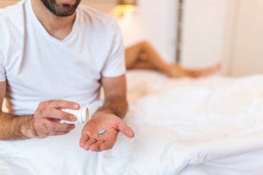 Mens health and sexual problems, male power and libido. Focus on blue pill in hands of happy young man, blurred smiling wife sits in bed in modern interior of bedroom, close up