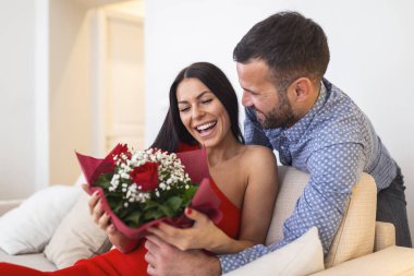 Excited young woman receiving unexpected Bouquet of red roses from husband at home, loving generous boyfriend making romantic surprise to attractive girlfriend on Valentines day occasion