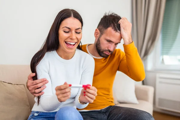 Not ready, worried man checking a pregnancy test with his excited wife sitting on a couch at home