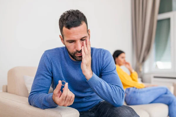 Stressed man holding her wedding ring worried about breakup or divorce, avoid talking with wife after fight, anxious couple ignore each other. Frustrated sad husband taking off the wedding ring