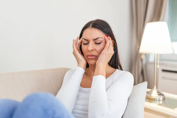 Portrait of an attractive woman sitting on a sofa at home with a headache, feeling pain and with an expression of being unwell. Upset depressed woman lying on couch feeling strong headache migraine.