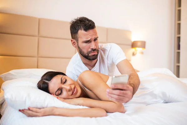 Jealous husband spying the phone of his partner while she is sleeping in a bed at home. Shocked jealous husband spying the phone of his wife while she sleeping in bed at home