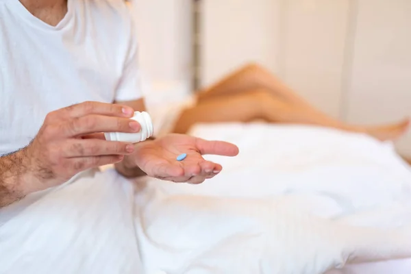 Mens health and sexual problems, male power and libido. Focus on blue pill in hands of happy young man, blurred smiling wife sits in bed in modern interior of bedroom, close up