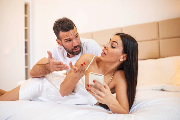 Photo of young couplein bed, woman use smartphone try explain envy angry husband she dont have another man point hand screen feel confused. Jealous husband spying his wife mobile phone