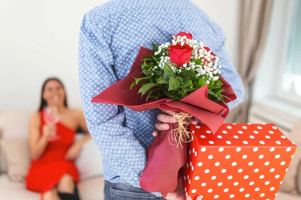 Unexpected moment in routine everyday life Cropped photo of man\'s hands hiding holding chic bouquet of red roses and gift box behind back, happy woman is on blurred background