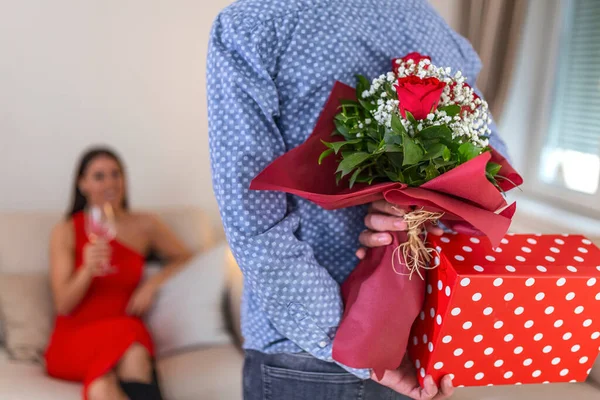 Unexpected moment in routine everyday life! Cropped photo of man\'s hands hiding holding chic bouquet of red roses and gift box behind back, happy woman is on blurred background