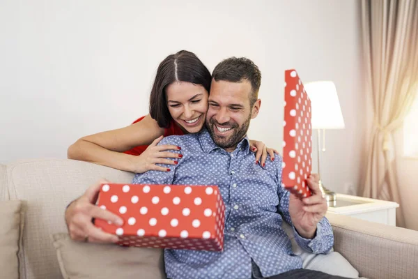 Woman surprising her husband with a gift on the couch at home on Valentine\'s Day. The beautiful young couple is celebrating at home. A woman is giving her man a gift box