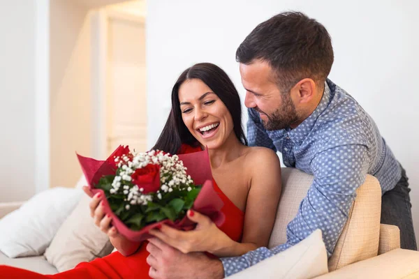 Excited young woman receiving unexpected Bouquet of red roses from husband at home, loving generous boyfriend making romantic surprise to attractive girlfriend on Valentines day occasion