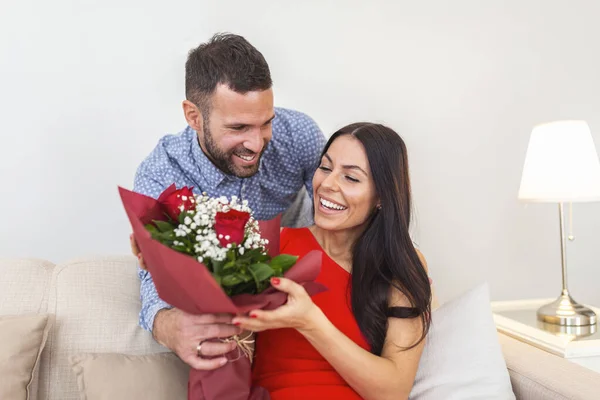 Excited young woman receiving unexpected Bouquet of red roses from husband at home, loving generous boyfriend making romantic surprise to attractive girlfriend on Valentines day occasion