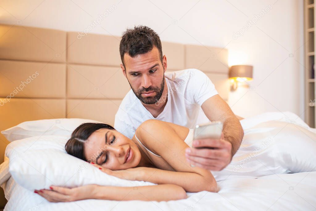 Jealous husband spying the phone of his partner while she is sleeping in a bed at home. Shocked jealous husband spying the phone of his wife while she sleeping in bed at home