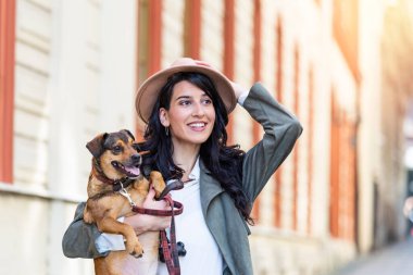 Sokakta yürürken şapkalı, ütülü ve köpeğe sarılan genç bir kadın. Gülümseyen genç bir kadın güzel bir günün tadını çıkarıyor ve evcil hayvanla poz veriyor. Köpeğiyle oynayan bir kadın.