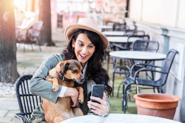 Köpeğiyle birlikte evcil hayvan kafeteryasında genç bir kadın. Yerel bir kafede köpeğiyle kahve içen bir kız cep telefonuyla selfie çekiyor.